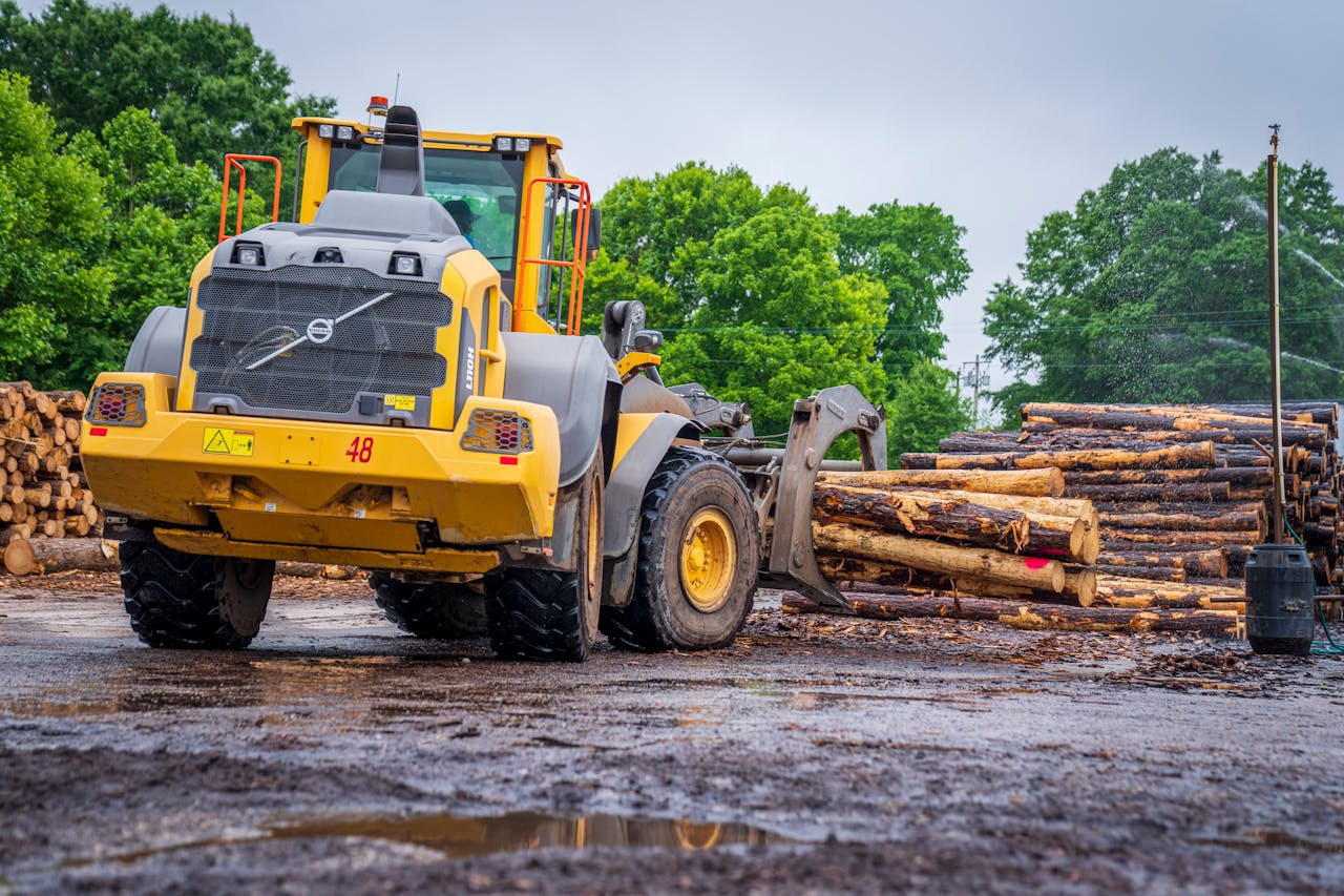 Yellow industrial loader carrying tree logs in a muddy lumber yard setting.